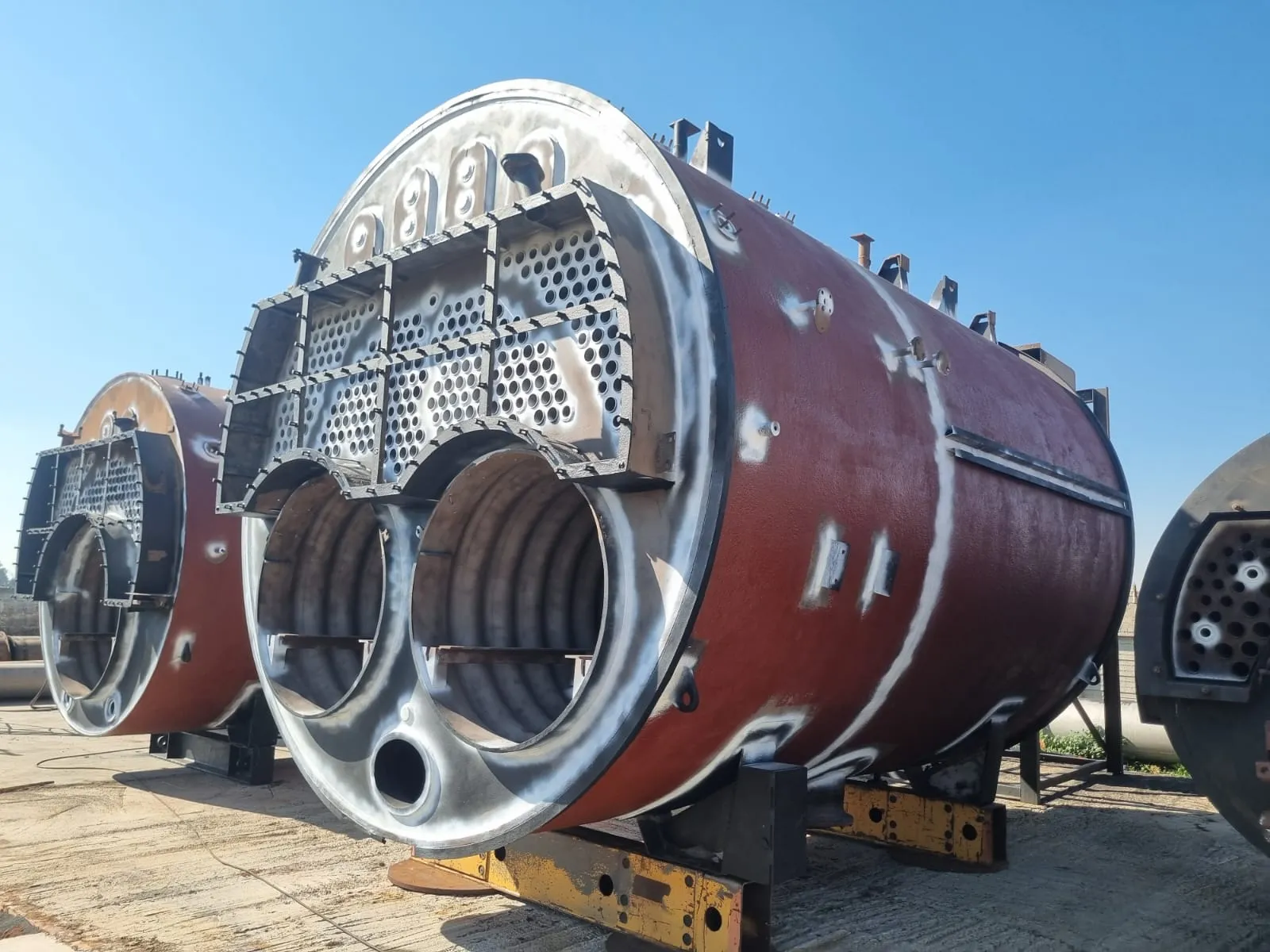 Three cylindrical boiler shells standing on steel supports in an open yard. The nearest has its tube plate drilled and its corrugated furnace tubes open to view, with red oxide primer across the barrel