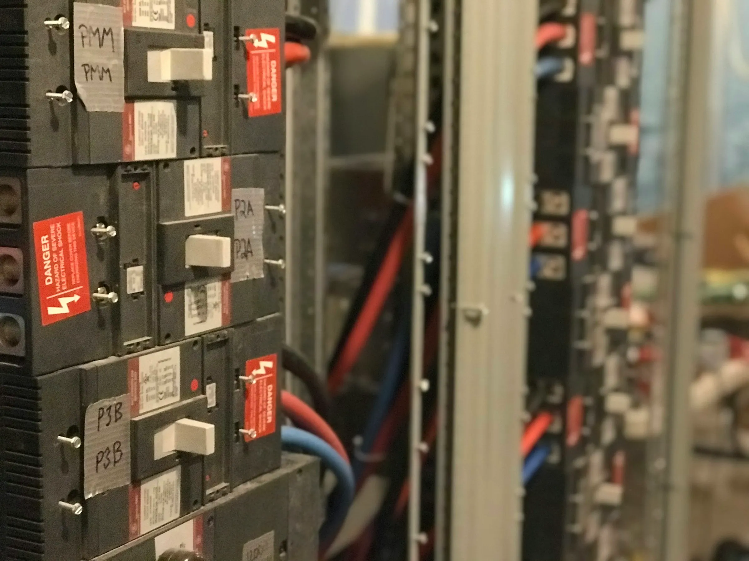 Interior of an electrical distribution board — rows of moulded-case circuit breakers with handwritten circuit labels, red electric-shock warning notices, and red and blue cabling running behind them
