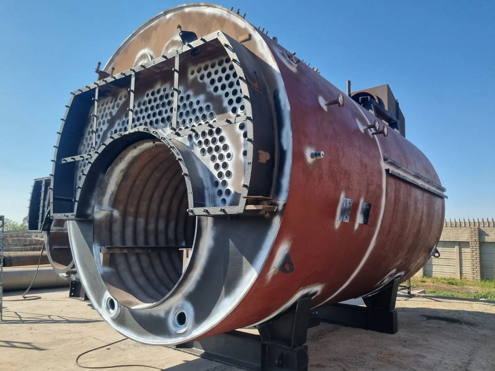 The open end of a cylindrical steel shell: a drilled tube plate, the corrugated furnace tube behind it, welded stiffening brackets, and red oxide primer across the outside.