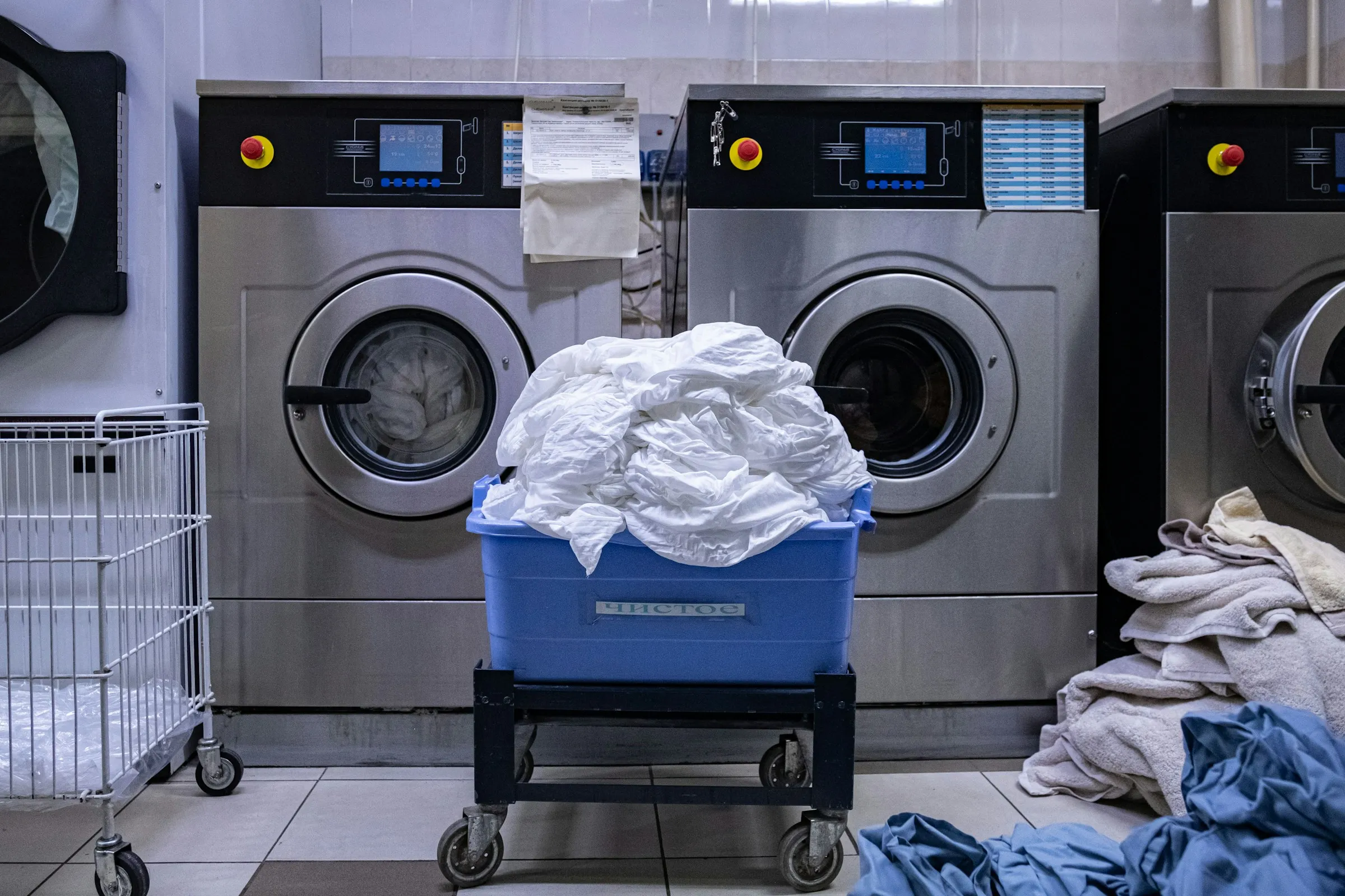 Commercial washer-extractors with industrial control panels, a linen trolley loaded with white linen in front of them