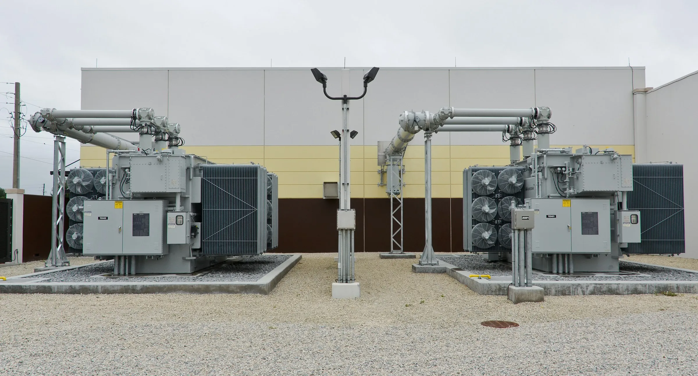 Two outdoor power transformers on concrete plinths beside a building, with radiator banks, bushings and overhead connections