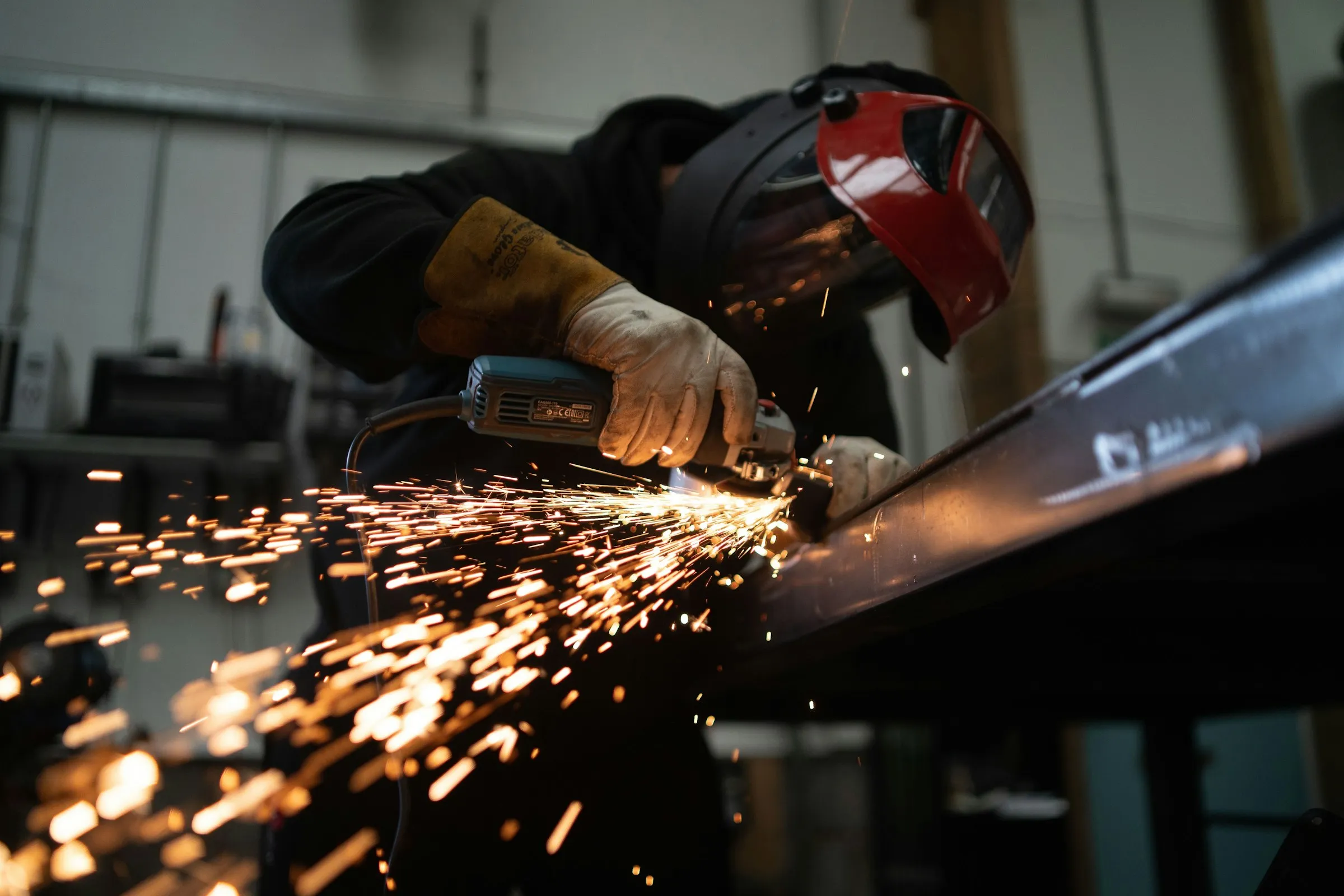 A person in a welding helmet and leather gauntlets running an angle grinder along a steel beam in a workshop, with a spray of sparks across the frame